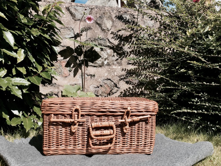 Wicker picnic basket sitting on a rug in front of a stone wall and near a pink flower.