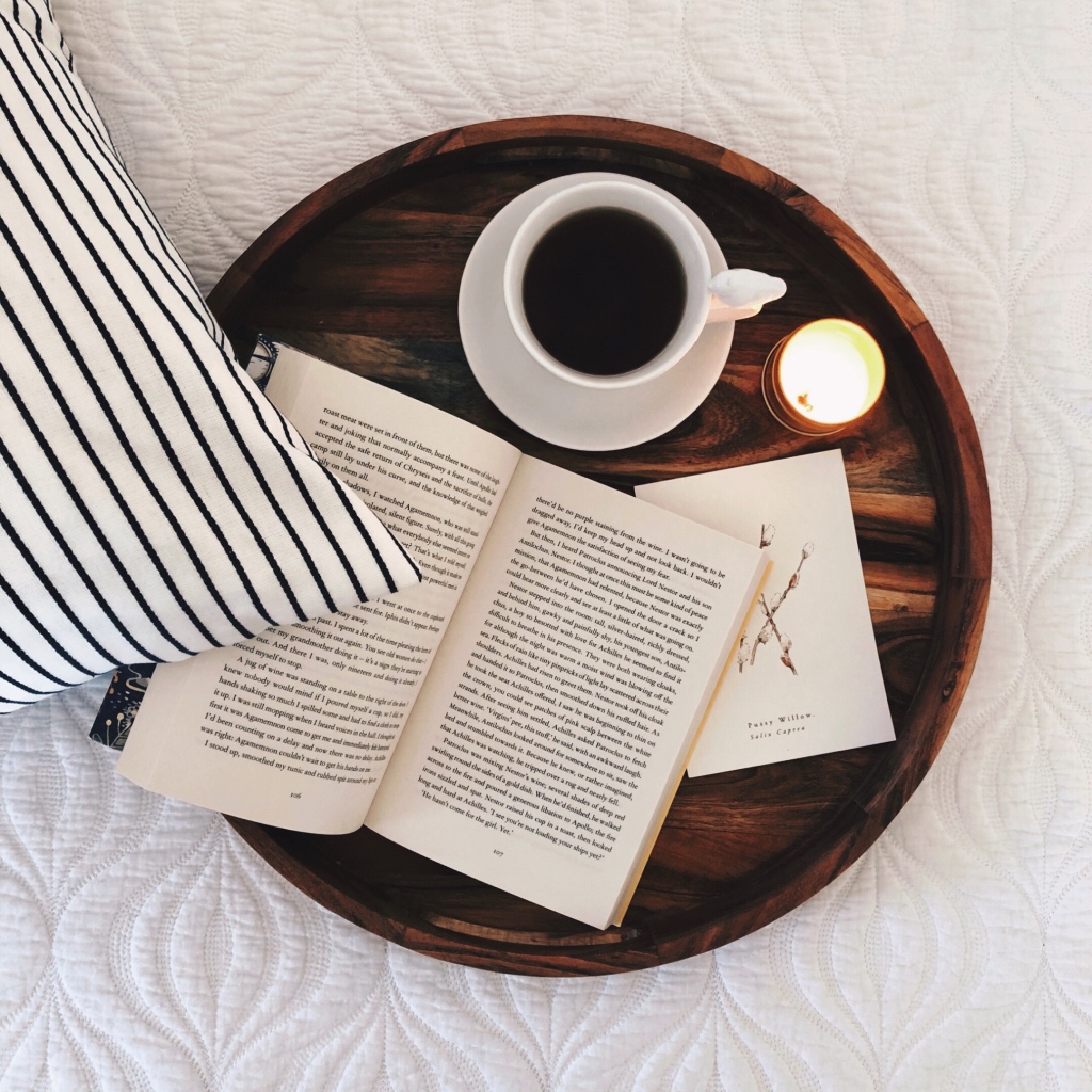 Book, candle, tea cup and small print on a round timber tray set on a white coverlet beside a blue and white stripe cushion.