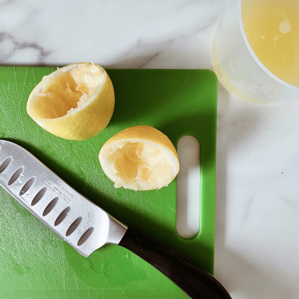 Juiced lemon halves on a green chopping board with a knife, beside a jug of lemon juice.