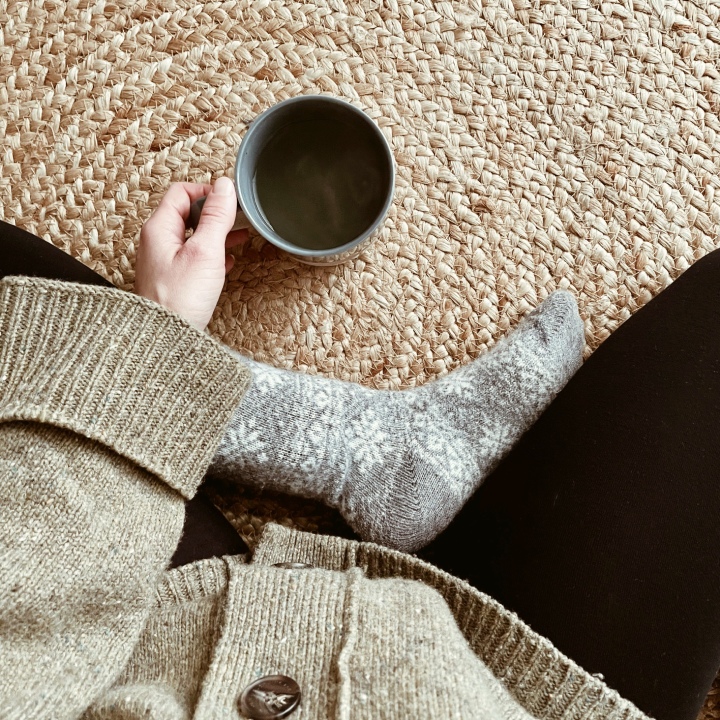 Looking down on a woman in an oversized olive green cardigan, wearing grey fair isle socks and black leggings, holding a cup of steaming tea on a jute rug.