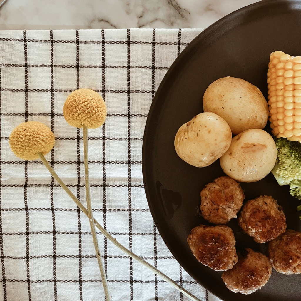 Plate of Swedish meatballs, broccoli, corn and potatoes atop a black and white checked tablecloth besides two Billy Button stems.